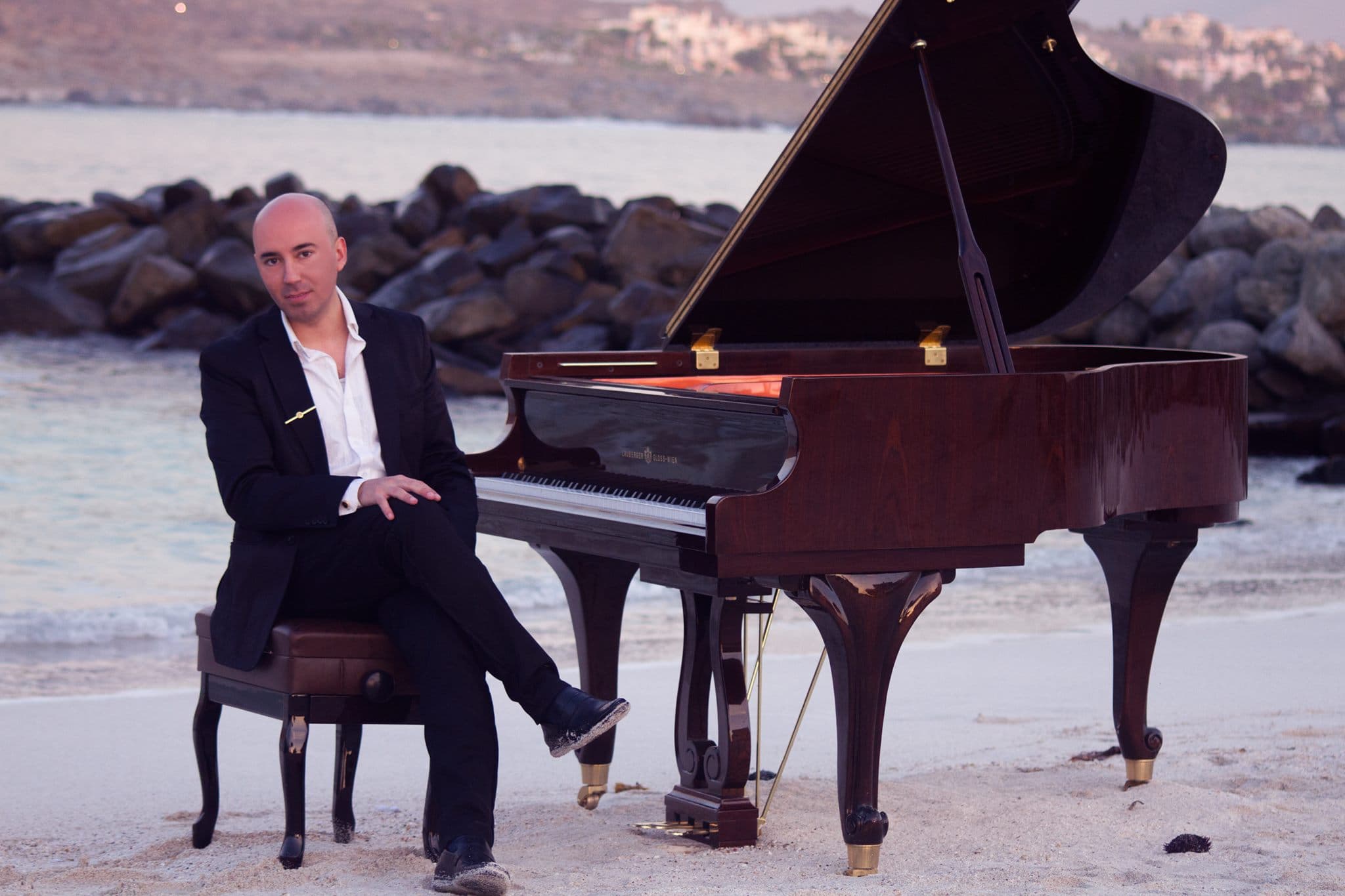 Juan Estay sitting at a grand piano on the beach with ocean and rocks in the background